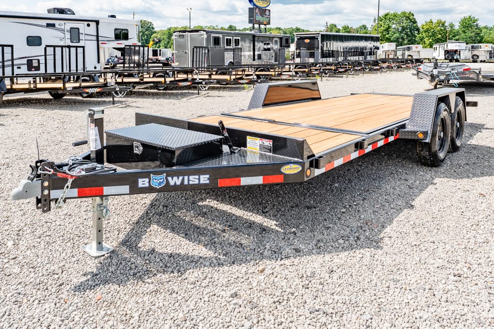 Flatbed car trailer with a wooden deck and dual axles on a gravel lot, brand name Wise on the frame, with other trailers in the background.