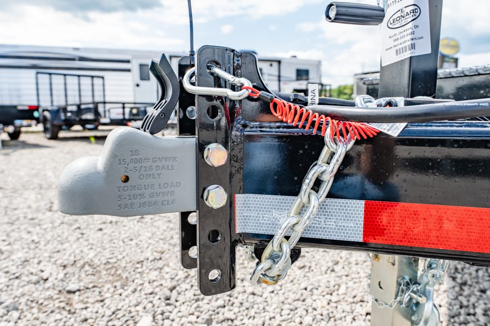 Trailer hitch with a safety chain, lock, and reflective tape on a gravel lot; RVs in the background.