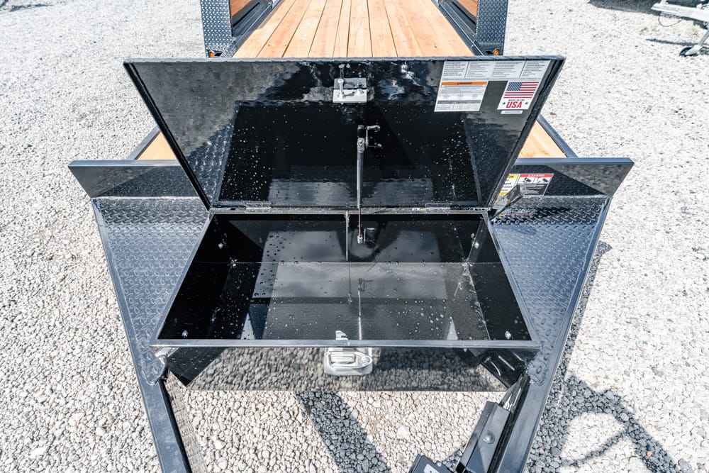 Open black metal storage toolbox on a utility trailer with diamond-plate sides, resting on a gravel lot; interior appears empty.
