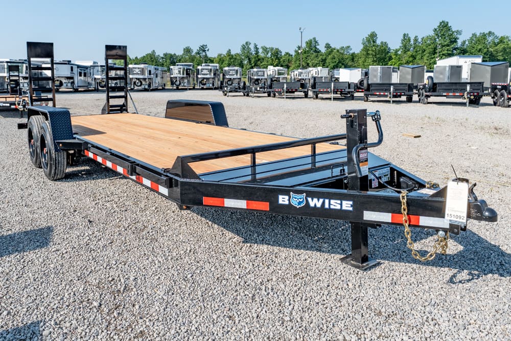 Black B-WISE flatbed trailer with a wooden deck on a gravel lot, others trailers visible in the background at a trailer lot park.