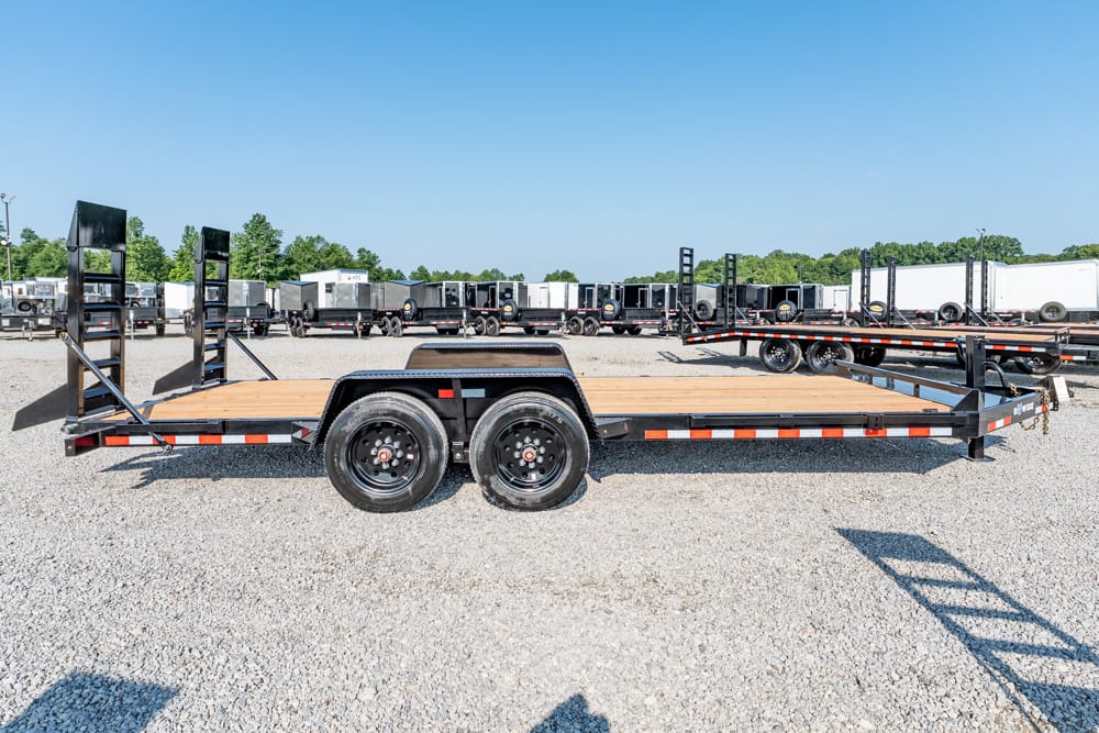 Dual-axle flatbed trailer with a wooden deck and foldable ramps at the rear, parked on a gravel lot among other trailers.
