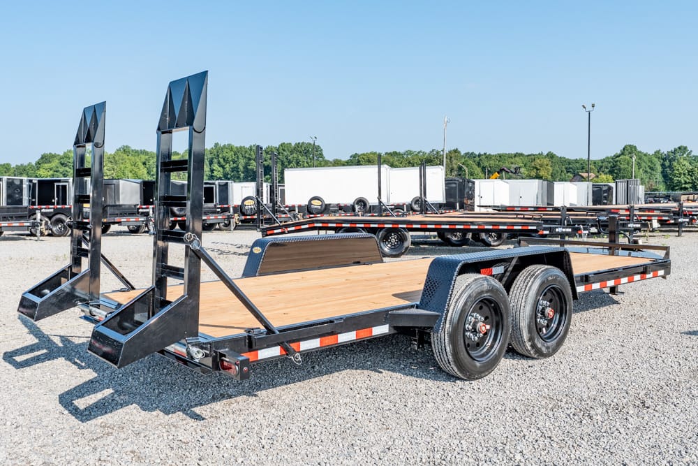 Flatbed trailer with dual axles and fold-down ramps in a gravel lot of trailers, ready to haul equipment.