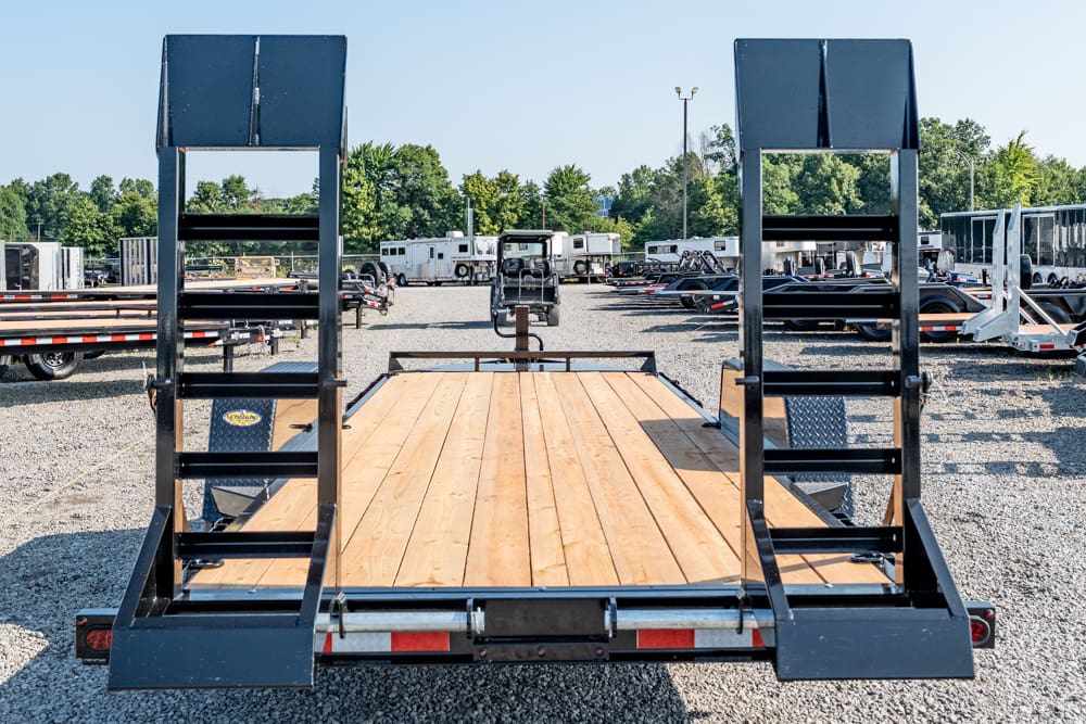 Rear view of a wooden flatbed trailer with metal ramps folded upright, in a gravel lot with other trailers nearby.