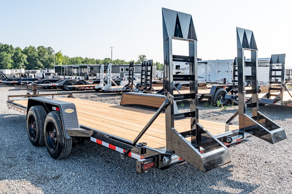 Dual-axle flatbed trailer with a wooden deck and fold-down loading ramps in a gravel lot.