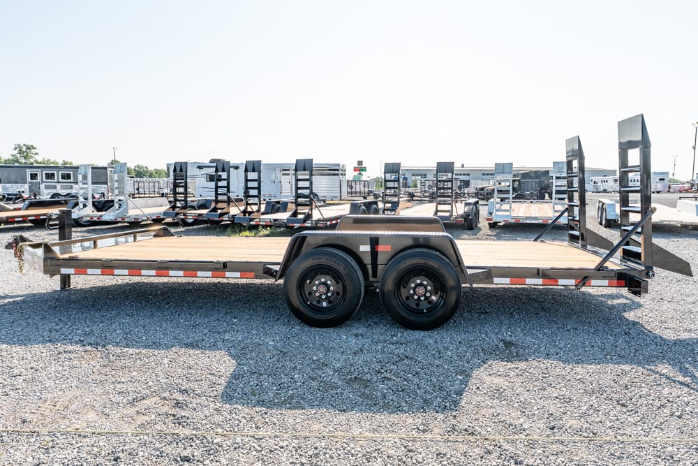 Flatbed trailer with dual axles and loading ramps, parked on a gravel lot with other trailers in the background.