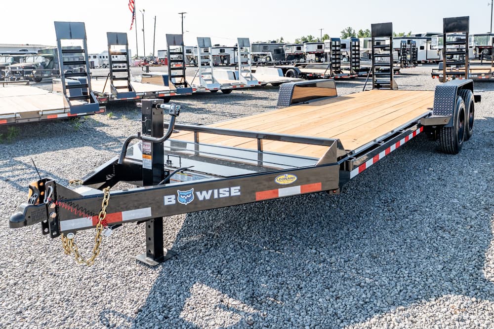 Black steel flatbed trailer with a wooden deck parked in a gravel lot among other trailers at a dealership.