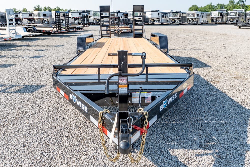 Front view of a black flatbed trailer with a wooden deck and safety chains, in a gravel lot with other trailers behind it.
