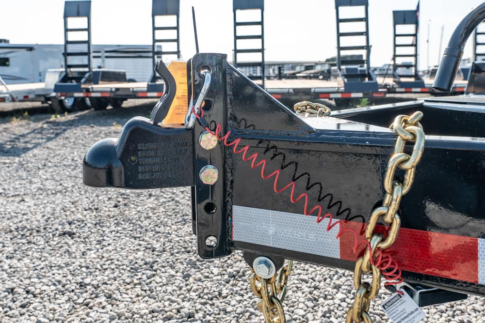 Close-up of a trailer hitch with safety chains and a red spring latch on a gravel lot.