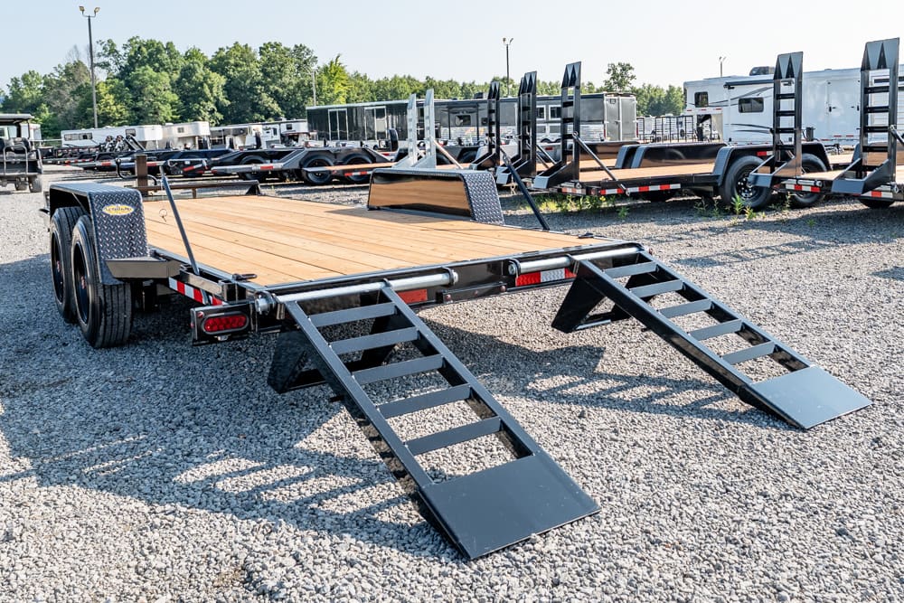 Flatbed trailer with two fold-down ramps parked on a gravel lot among other trailers.