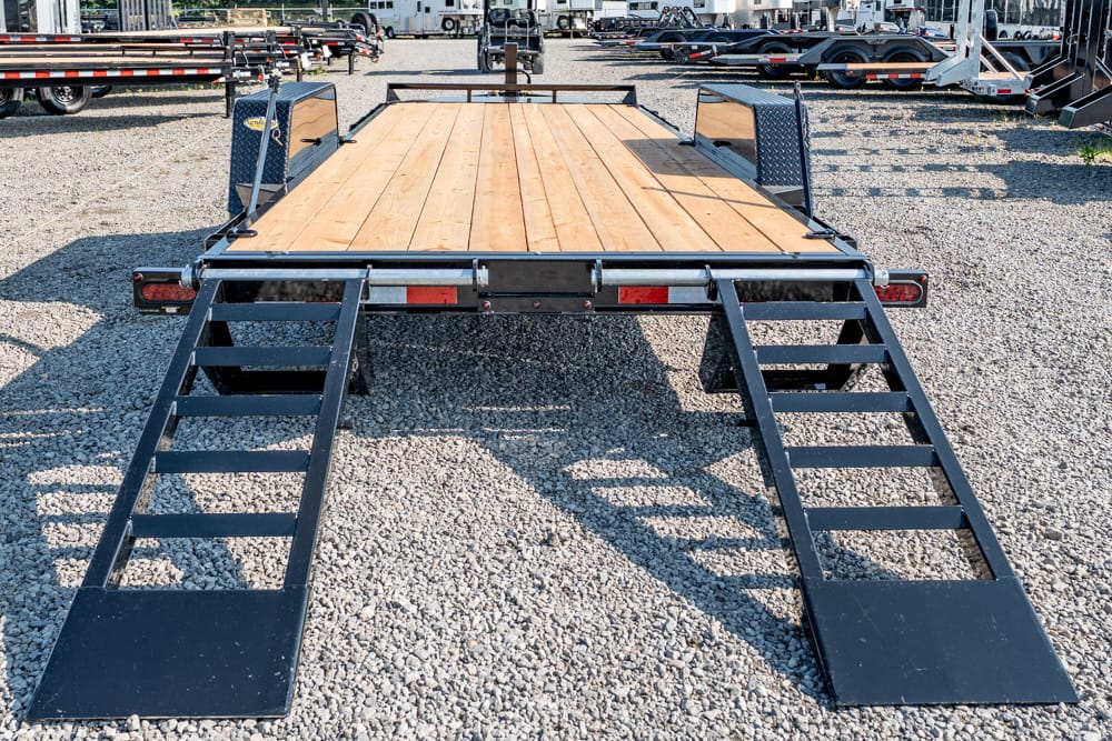 Black metal trailer ramps folded down from a wooden flatbed trailer on a gravel lot, with other trailers visible in the background.