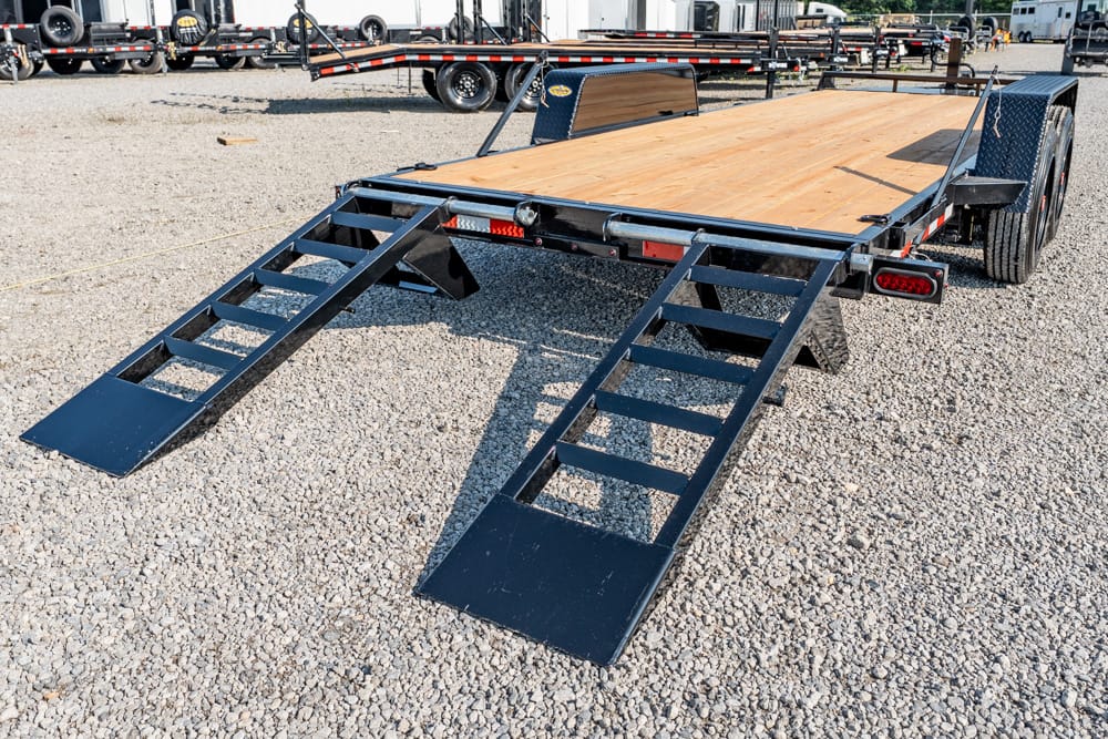 Flatbed trailer with wooden deck and two metal loading ramps extended on a gravel lot at the rear.