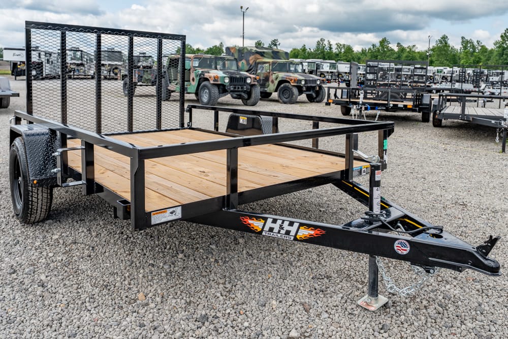 Flatbed utility trailer with wooden deck and side rails, hitched to a tow, parked on a gravel lot with other trailers nearby.