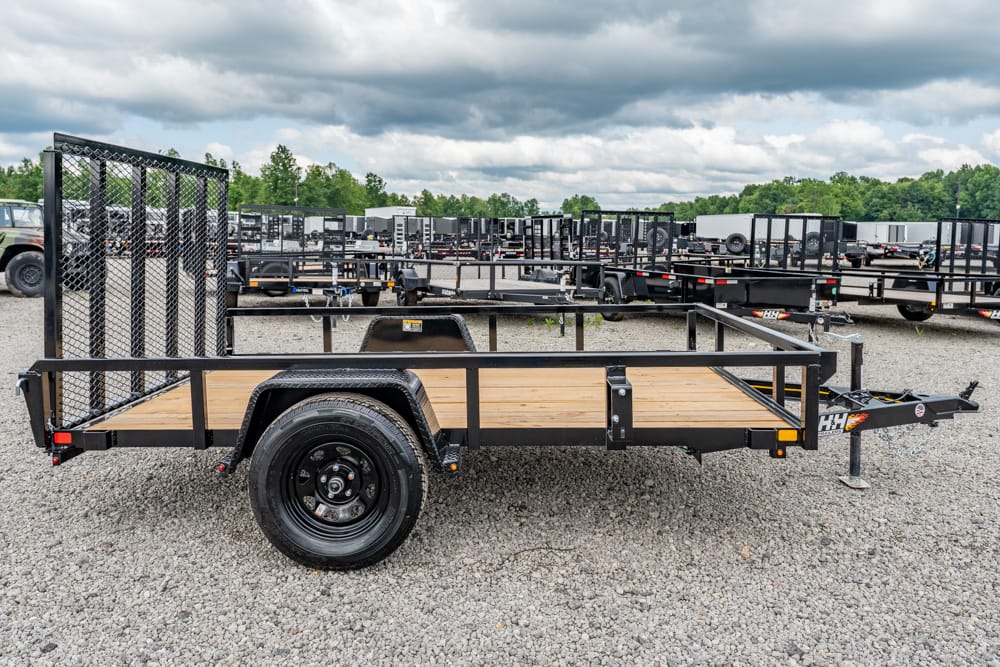 Black utility trailer with a wooden deck and rear mesh gate on a gravel lot.