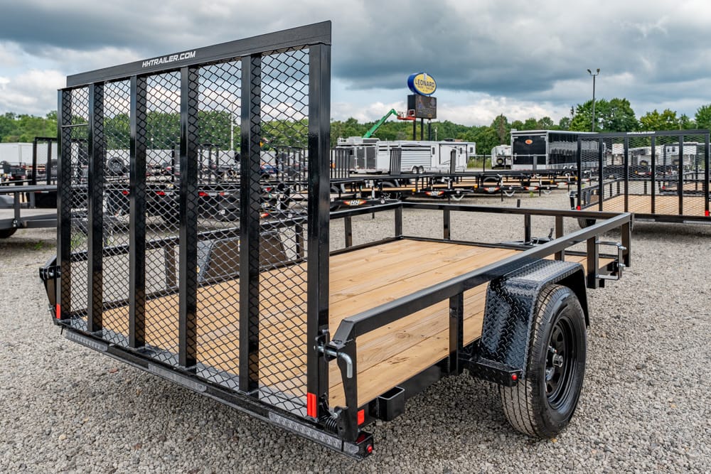 Open utility trailer with a wooden deck, black metal mesh tailgate, and single axle on a gravel lot.