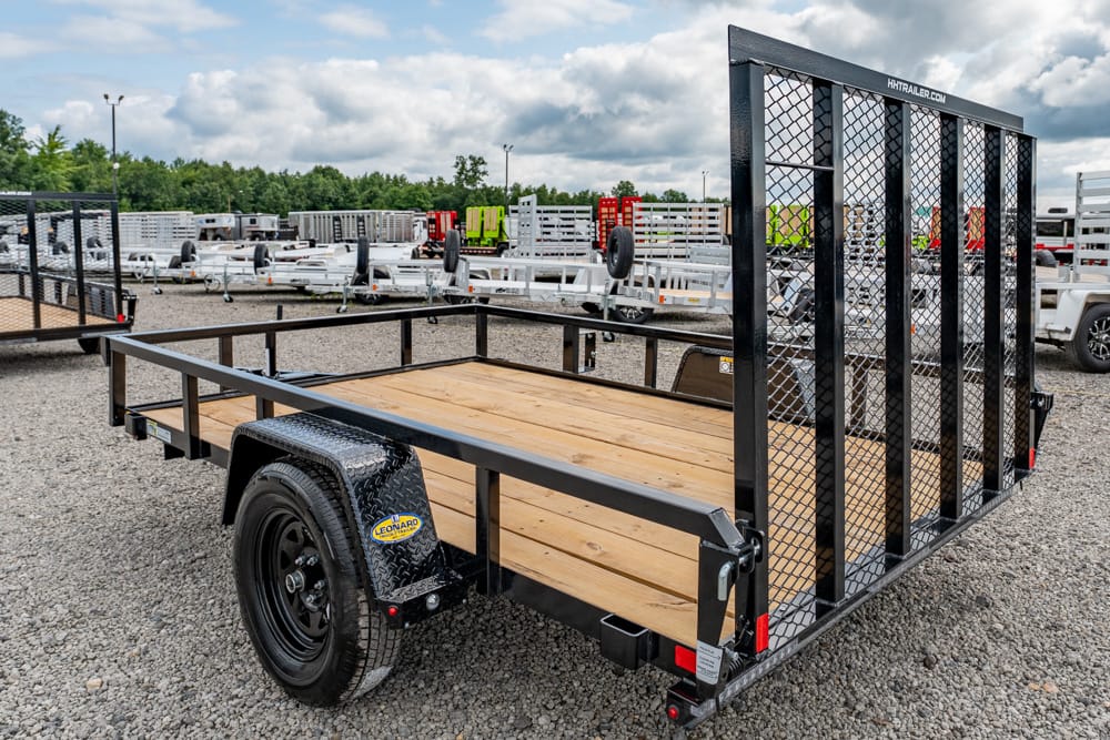 Flatbed utility trailer with a wooden deck and a tall mesh ramp at the rear, parked on a gravel lot with other trailers in the background.