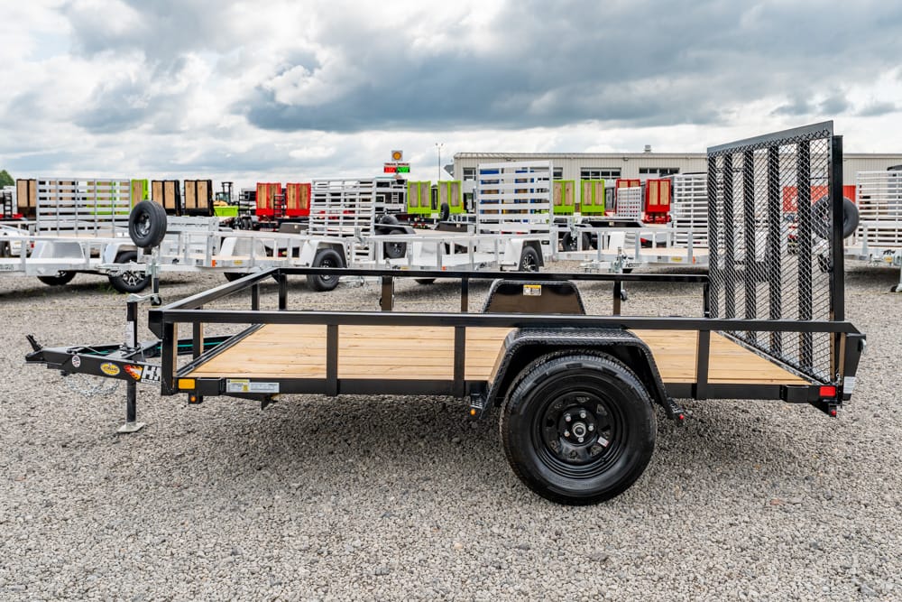 Flatbed utility trailer with a wooden deck and tall black side rails parked on a gravel lot, surrounded by other trailers in a dealer yard.
