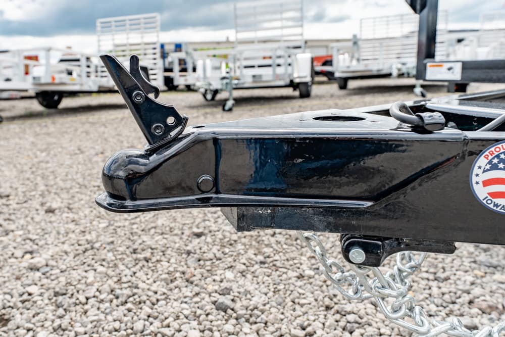 Close-up of a black trailer hitch with a curved latch and safety chains on a gravel lot, with other trailers in the background.