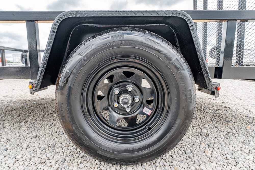 Close-up of a trailer wheel: black steel rim and tire, with a black fender above, resting on a gravel surface.