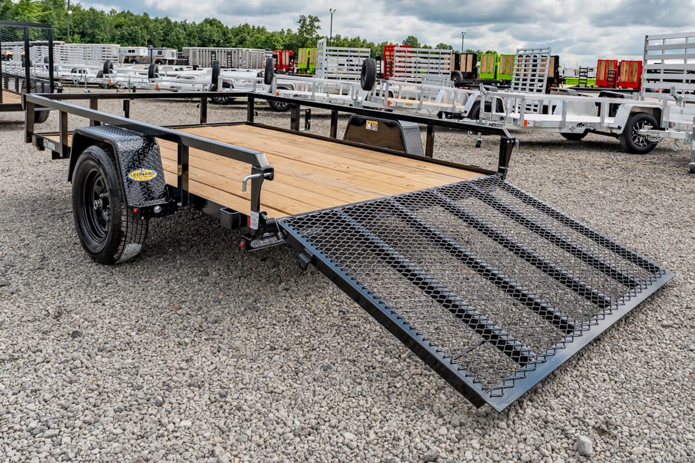 Flatbed trailer with a wooden deck and metal ramp lowered for loading on a gravel lot, with other trailers in the background.
