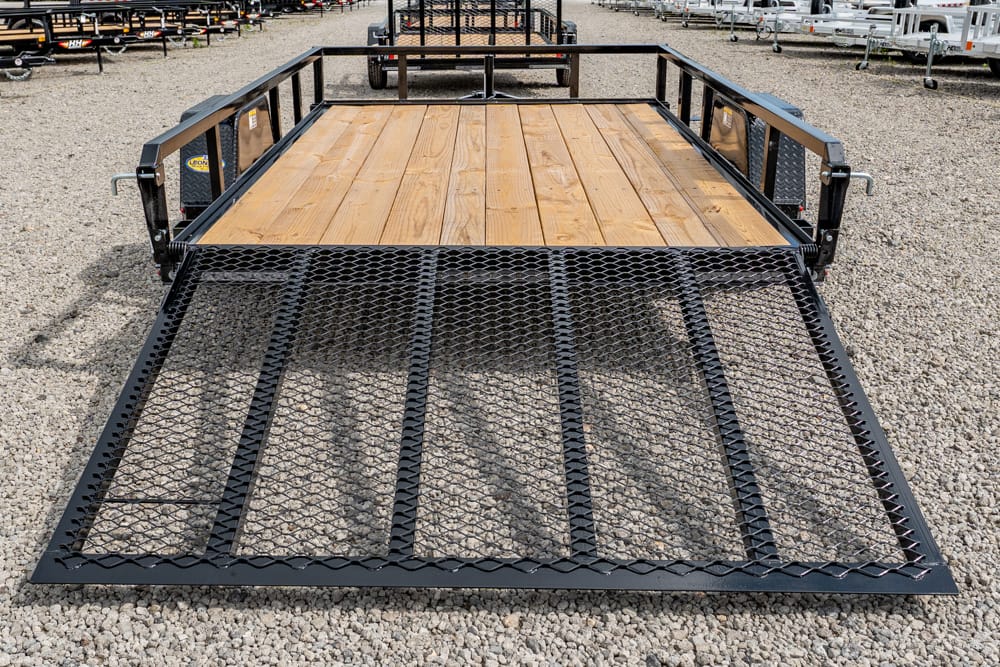 Flatbed trailer with a wooden deck and a metal mesh ramp lowered to the ground on a gravel lot, ready for loading.