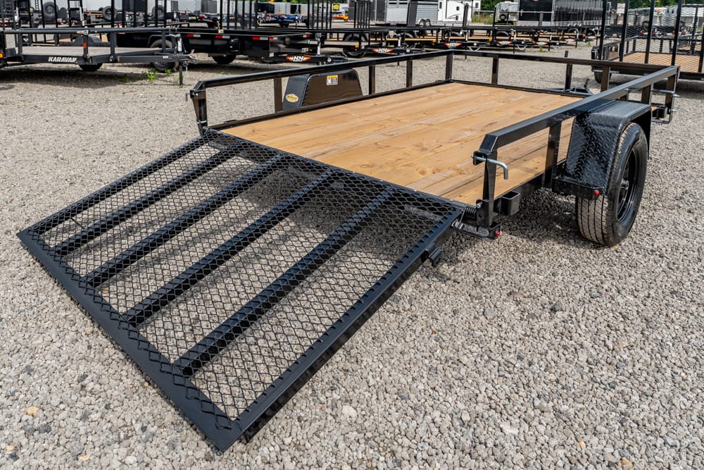 Black utility trailer with a wooden deck and a large fold-down mesh ramp on a gravel lot, spare tire mounted on the side.