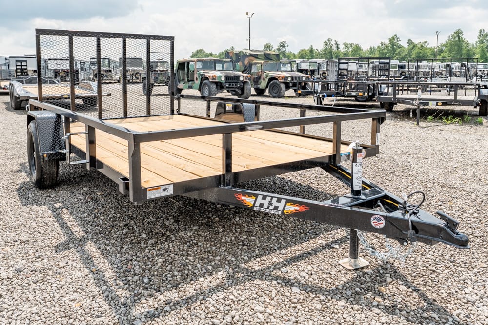 Flatbed utility trailer with wooden deck and dual axles in a gravel lot, surrounded by other trailers and military trucks in the background.