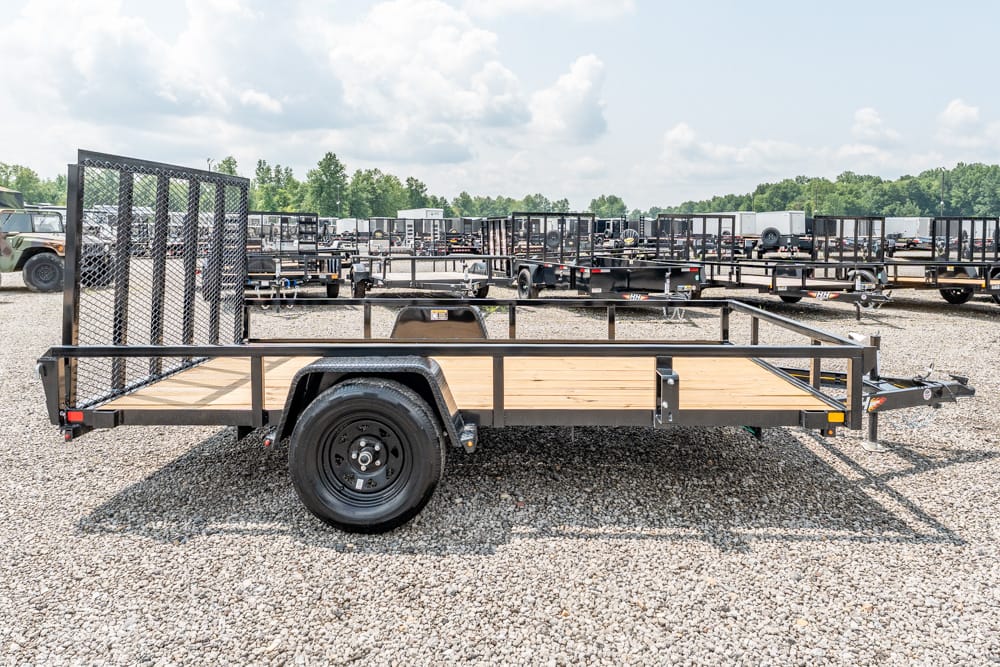 Flatbed utility trailer with a wooden deck and metal mesh gate, parked among other trailers in a gravel lot.