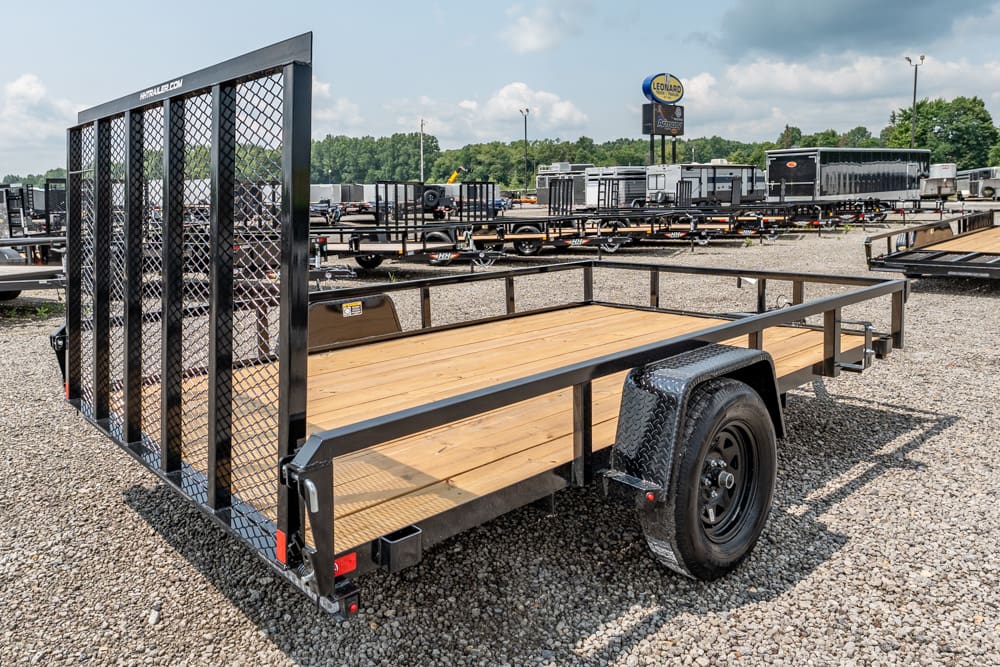 Flatbed trailer with a wooden deck and a tall metal ramp gate in a gravel lot filled with other trailers.