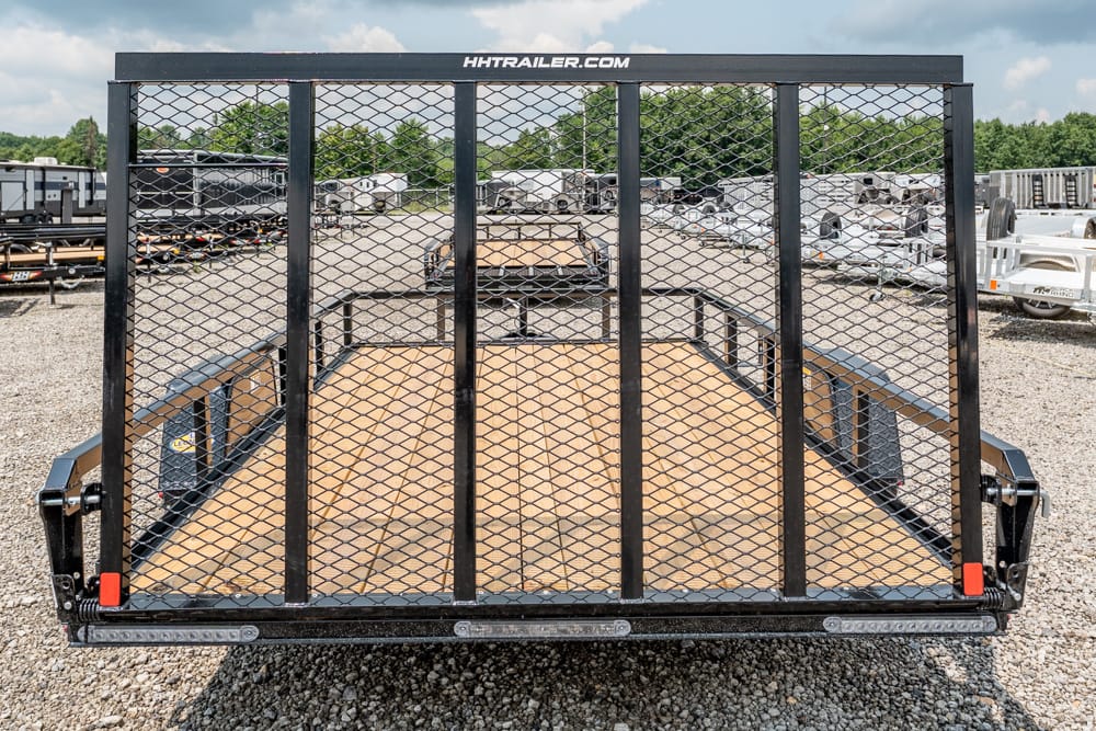 Rear view of a black utility trailer with a diamond-mesh ramp gate and wooden deck on a gravel lot.