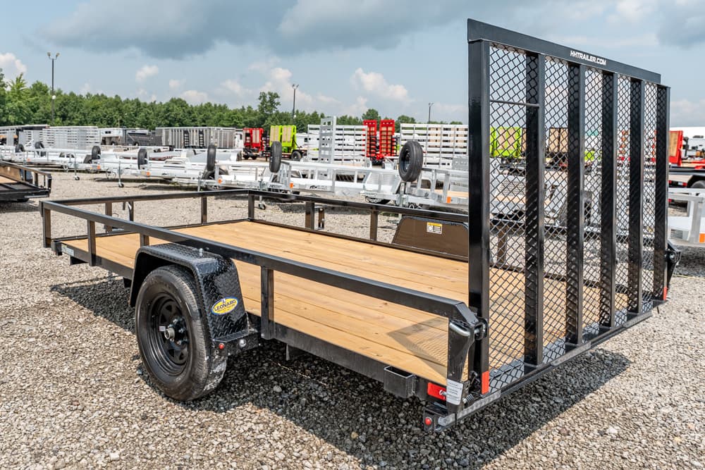 Flatbed trailer with wooden deck and a tall mesh ramp at the rear, parked on a gravel lot with other trailers in the background.