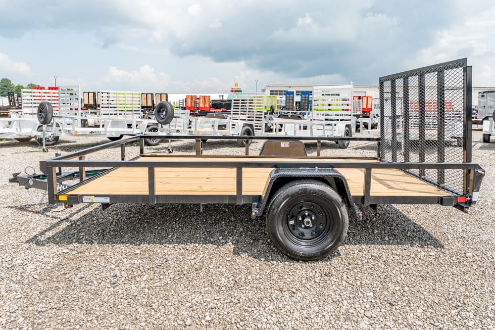 Utility flatbed trailer with wooden deck and side rails on a gravel lot, with a tall mesh ramp at the rear.