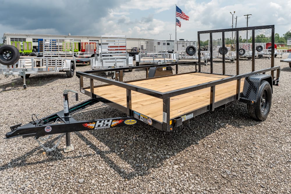 Black flatbed utility trailer with a wooden deck and a mesh rear gate on a gravel lot at a trailer yard.