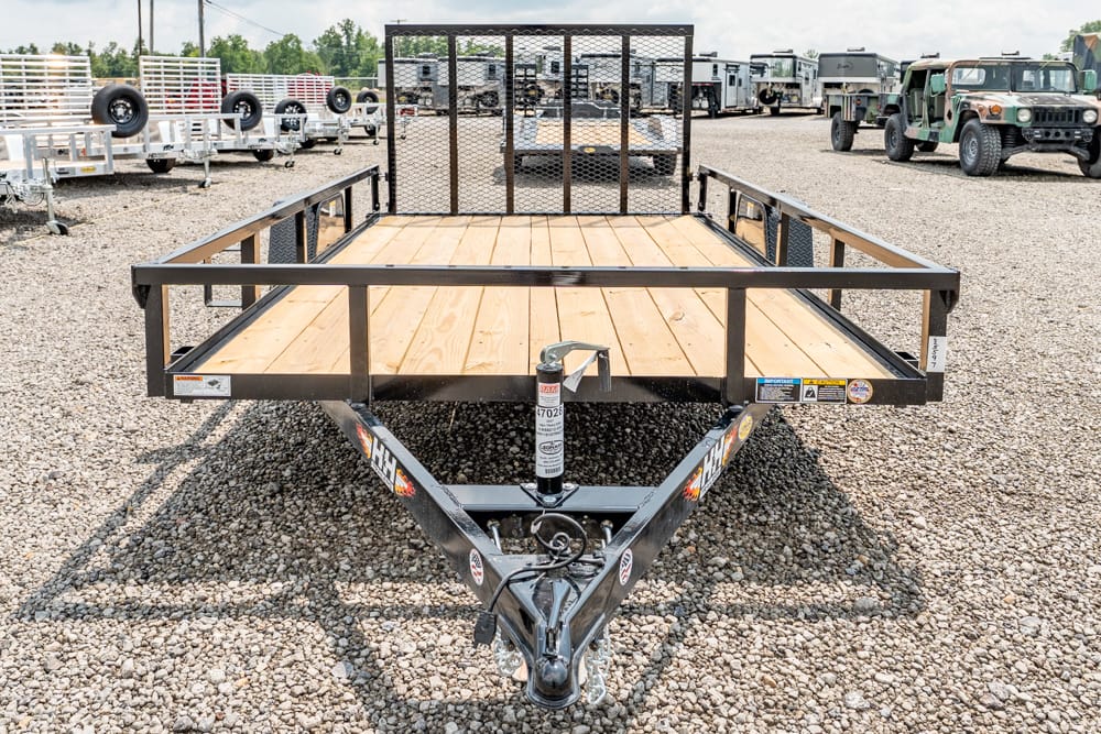 Flatbed utility trailer with a wooden deck and metal rails on a gravel lot, hitch and jack in the foreground.