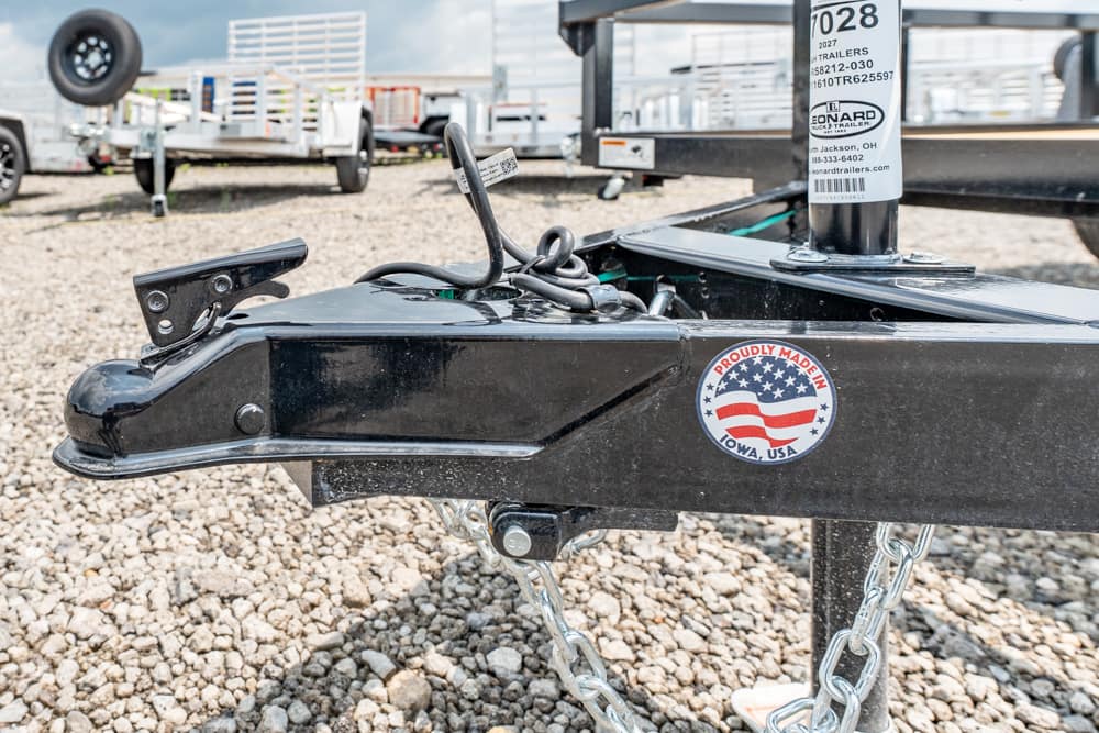 Close-up of a black trailer hitch with safety chains and an 'Iowa, USA' sticker, on a gravel lot with other trailers in the background