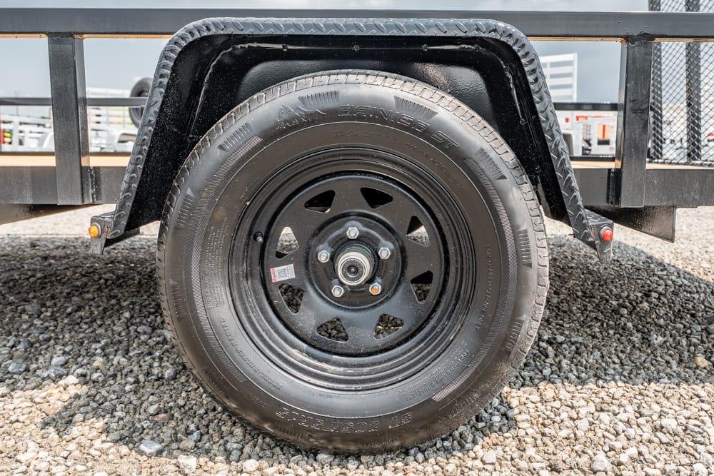 Trailer wheel with black steel rim and tire mounted under a metal fender on a gravel surface, utility trailer scene nearby