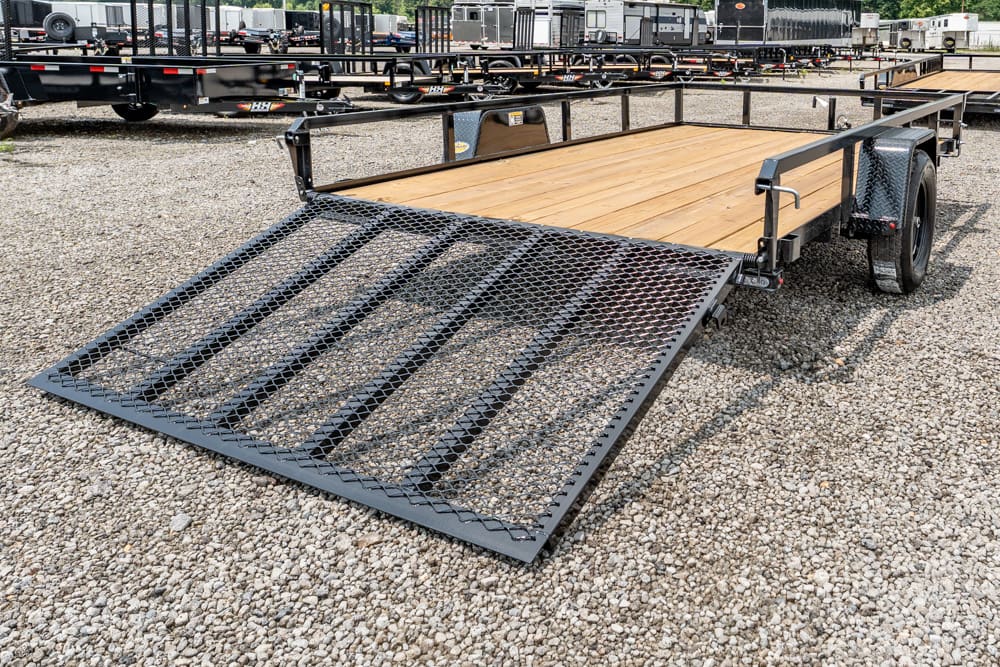 Flatbed trailer with a down-folding mesh ramp, wooden deck, parked on a gravel lot among other trailers." loading ramp down, ready for use.