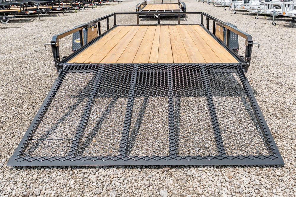 Black metal utility trailer with a wooden deck and diamond-mesh ramp in a gravel lot. The ramp is folded down, ready for loading.