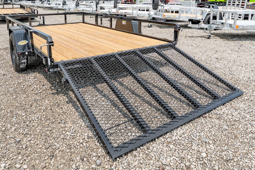 Flatbed utility trailer with a perforated metal loading ramp down on a gravel lot, wooden deck and other trailers in the background.