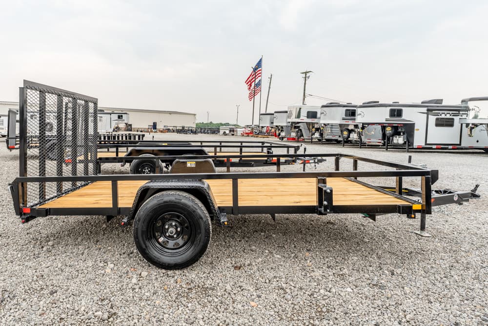 Flatbed utility trailer with wooden deck and black steel frame on a gravel lot, RVs and an American flag in the background.