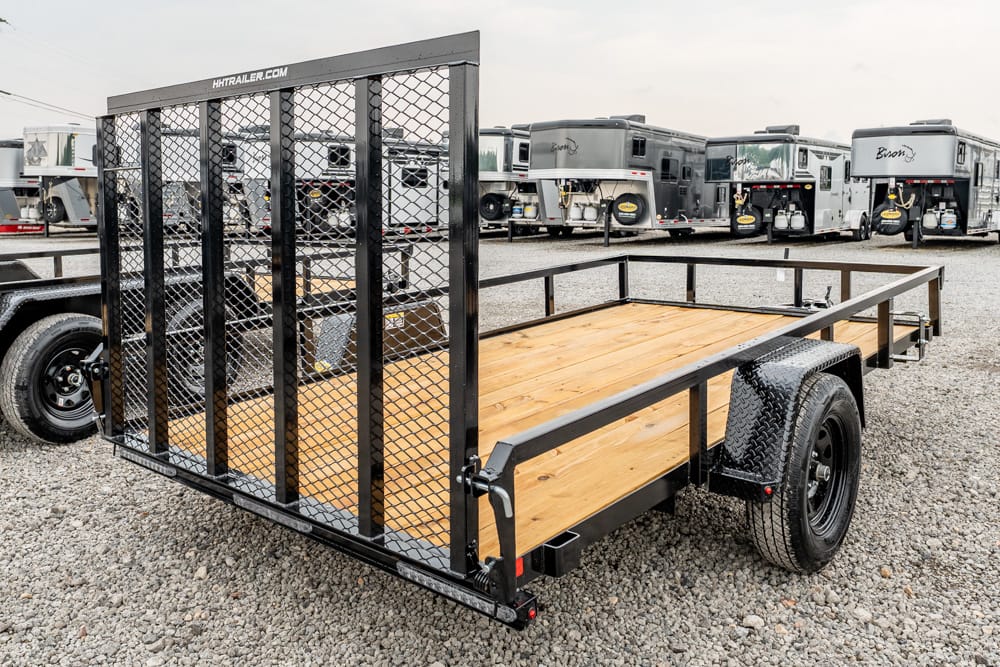 Utility flatbed trailer with a wooden deck and mesh ramp gate on a gravel lot; designed for hauling equipment or vehicles