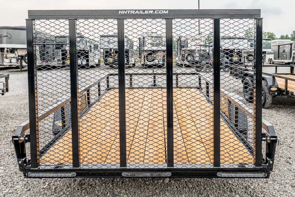 Rear view of a black utility trailer with a wooden deck and mesh ramp gate, parked among other trailers in a lot.