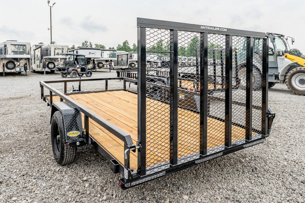Black metal utility trailer with a wooden deck and tall mesh gate parked on a gravel lot among RVs and construction equipment.