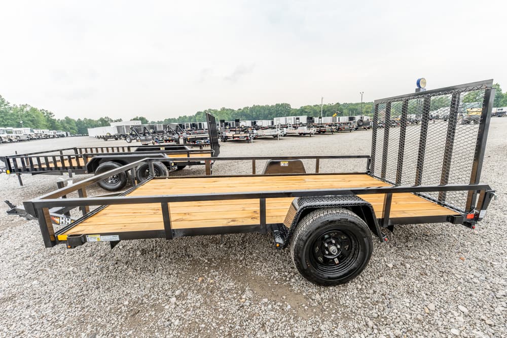 Flatbed utility trailer with a wooden deck and tall rear mesh ramp, parked on a gravel lot.