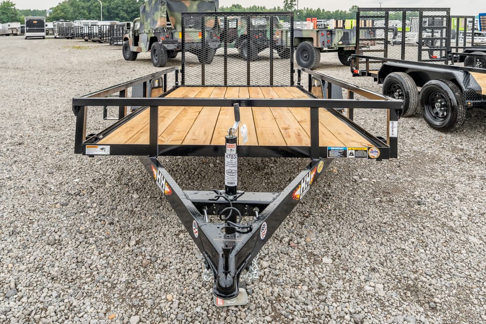 Front view of a black steel utility trailer with a wooden deck in a gravel lot, surrounded by other trailers and trucks.