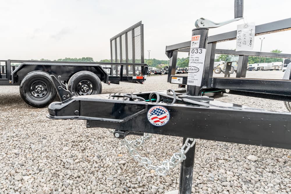 Close-up of a black trailer hitch with a safety chain on a gravel lot, dual-axle trailer visible in the background.