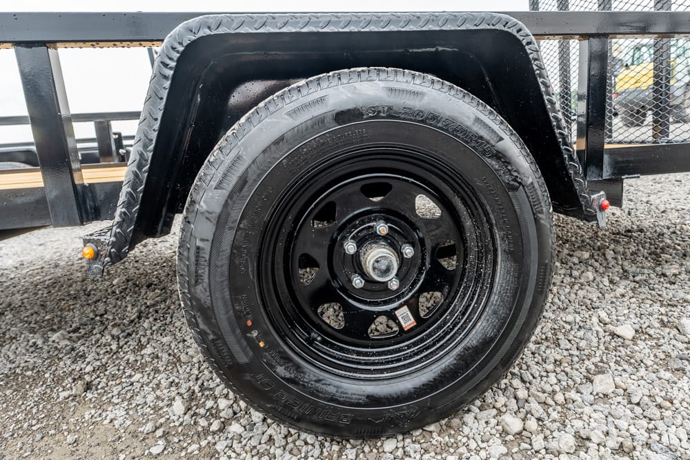 Trailer wheel with black steel rim and tire, mounted under a black fender on a gravel surface.