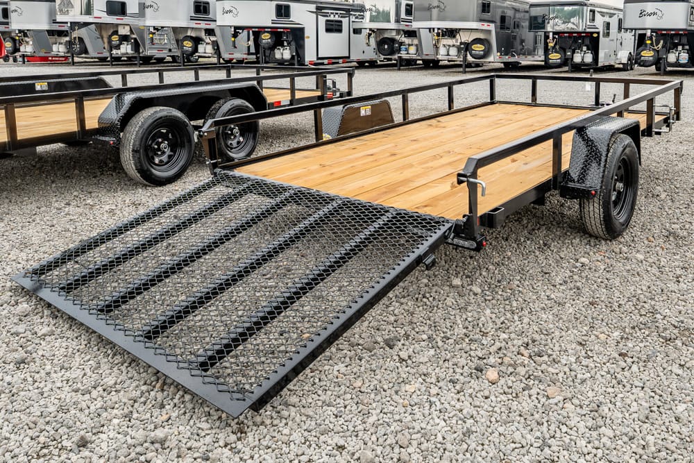 Flatbed trailer with a lowered metal mesh ramp and wooden deck on a gravel lot, with horse trailers in the background.