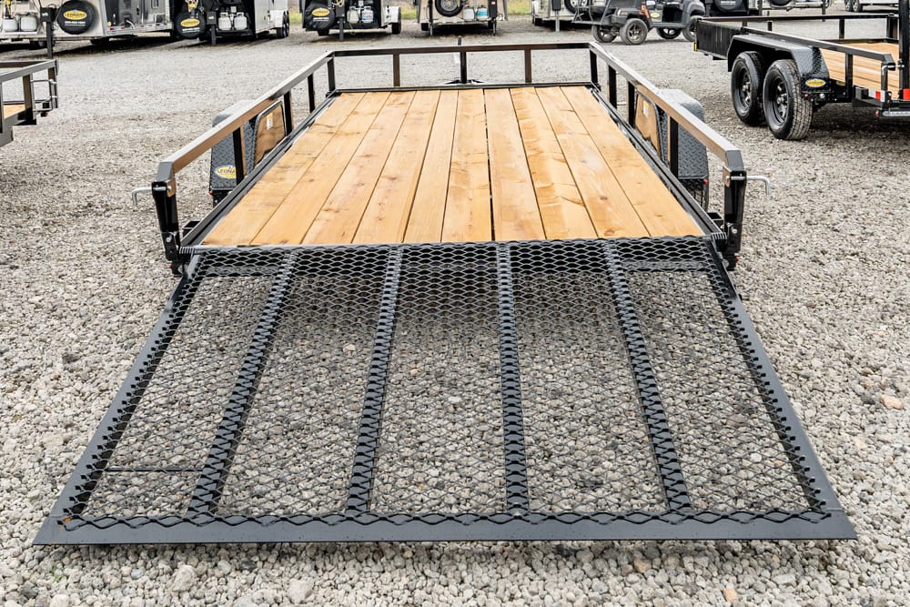 Flatbed trailer with wooden deck and a lowered metal ramp on a gravel lot, ready to load equipment