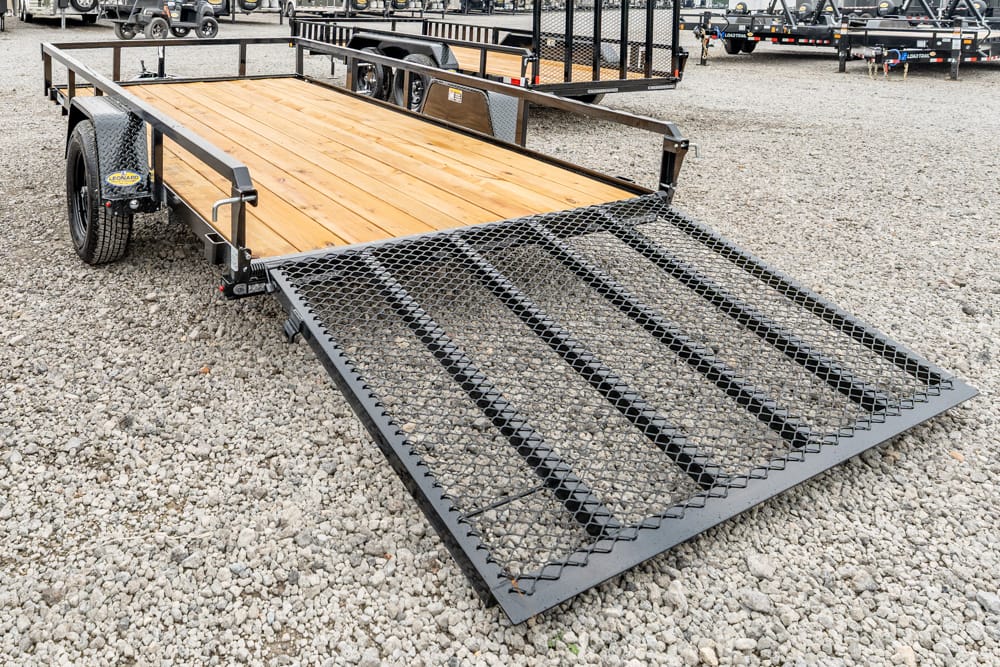 Flatbed trailer with a wooden deck and a heavy diamond-mesh ramp extended onto a gravel lot.