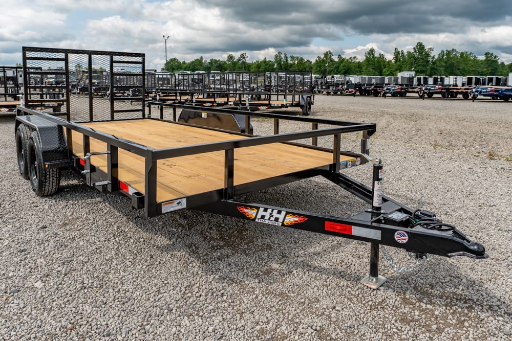Flatbed utility trailer with a wooden deck and black steel frame on a gravel lot, surrounded by other trailers in the background.